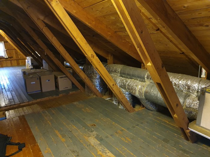 Isolerade ventilationsrör och ett FTX-aggregat monterat på en vind med lutande takstolar och kartonger på golvet.