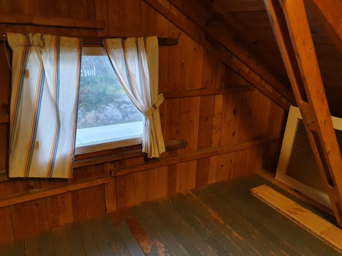 Vindrumsinteriör med träbeklädnad, ett fönster med gardiner och en lutande takkonstruktion.
