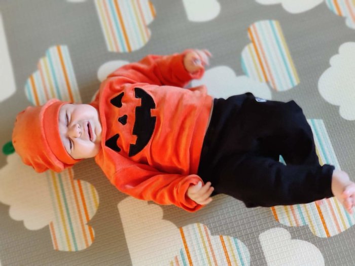 Bebis iklädd en pumpaliknande orange tröja med jack-o'-lanterntryck och svarta byxor, liggandes på en mönstrad matta.