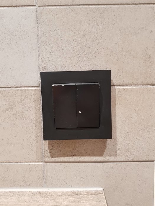 Dubbel ljusströmbrytare i svart på en vägg med beige kakel.