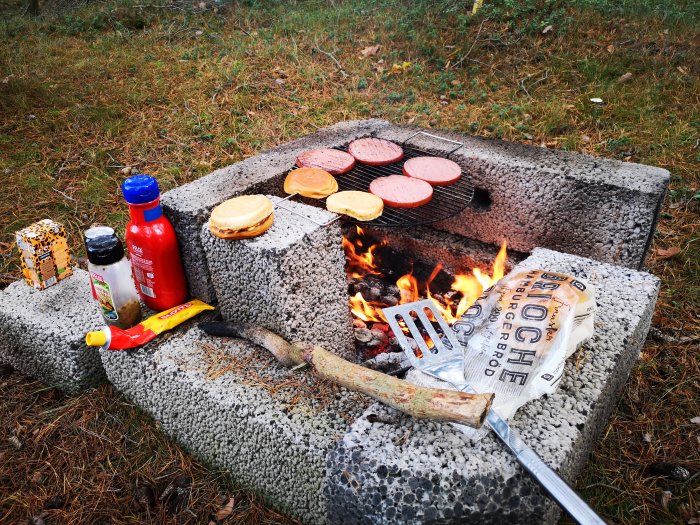 Hemmagjord grill av stenar med hamburgare som grillas och grilltillbehör runtomkring.