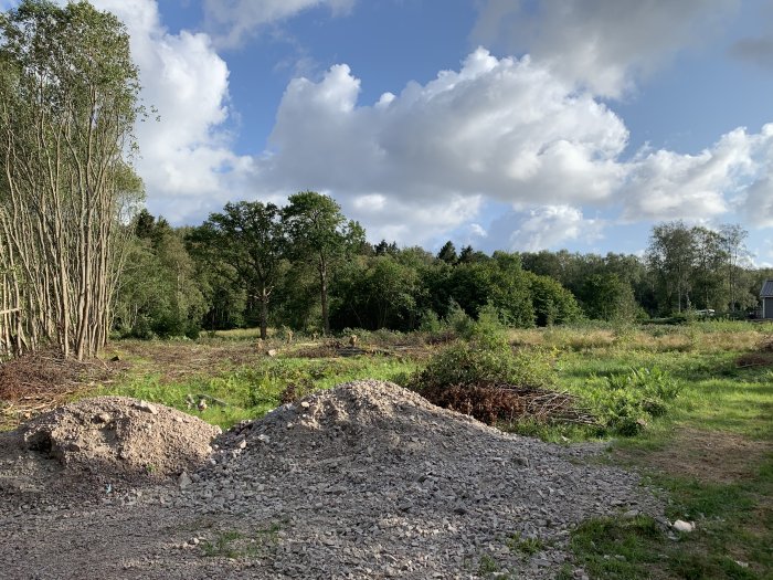 En rymlig infart med grushögar under en sommarhimmel, vid ett pågående byggprojekt i en grön omgivning.