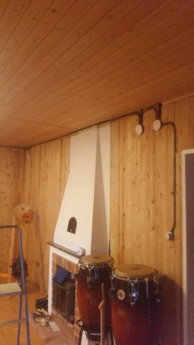 Vardagsrum under renovering med en oputsad spis, trumset och stege mot en vägg klädd med träpanel.