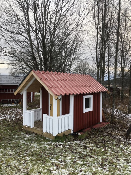 Röd lekstuga med nytt rött tak och vit veranda omgiven av gräs med lite snö.