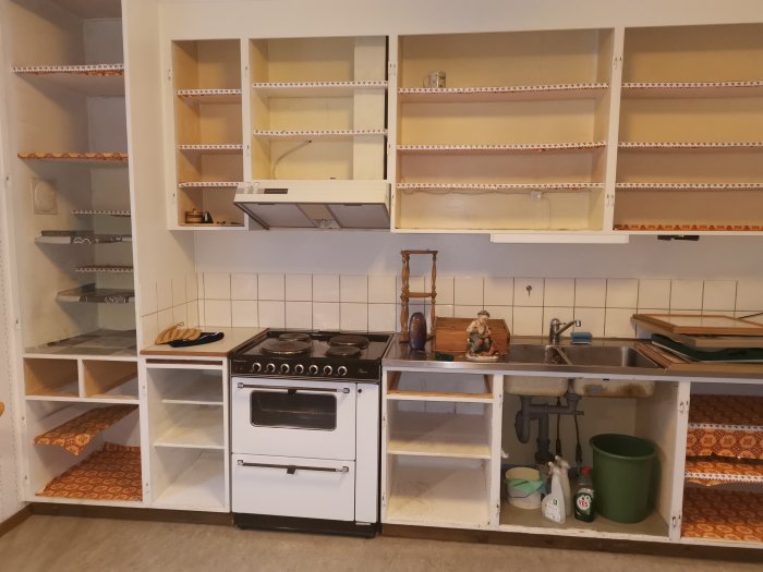 Kök före renovering med tomma hyllor, gammal spis och öppna skåpstommar.