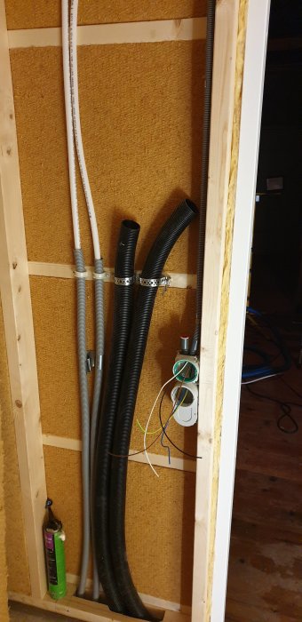 Öppen vägg med isolering där svarta och vita rör samt elledningar förekommer under förläggning.