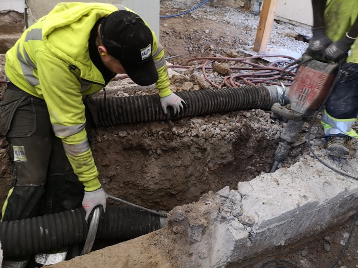 Arbetare använder handhållen såg för att skära igenom betong nära en dräneringssladd i en grävd grop.