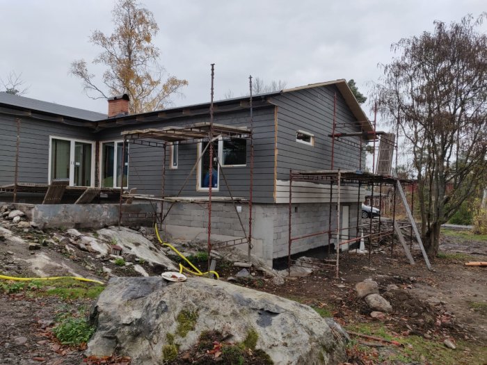 Hus under renovering med nymonterad panel och fönster, omgivet av byggställningar och ojämn terräng.