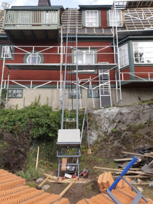 Ställning vid rött hus med vinschsystem på toppen för byggmaterial mot bergig terräng.