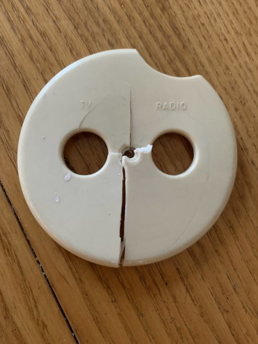 Sönderbrutet vitt lock för antennuttag märkt med "TV" och "RADIO", diameter 80 mm, ligger på träyta.