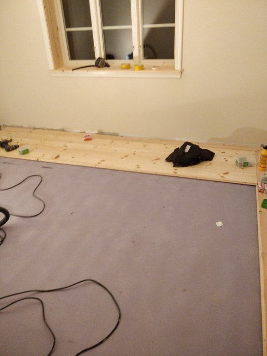 Påbörjat trägolv av furu längs med vägg, verktyg och materialet spred över skyddspapp på golvet.