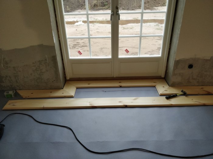 Pågående golvläggning med massiva furubrädor och verktyg nära ett fönster i ett rum under renovering.