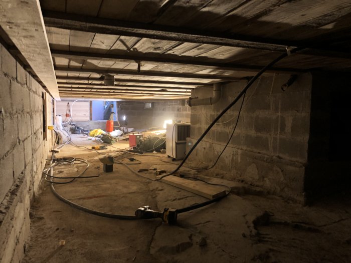 Dammtäckt källare under renovering med exponerade bjälkar och kablar längs golvet.