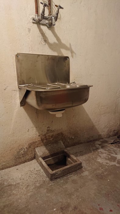 Väggmonterad vask ovanför en öppen spygatt i ett betonggolv i en källare.