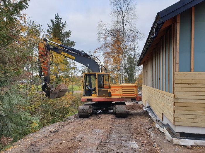 Grävmaskin flyttar fyllningsmaterial nära en nybyggd träbeklädd husfasad och ett trädrikt landskap.