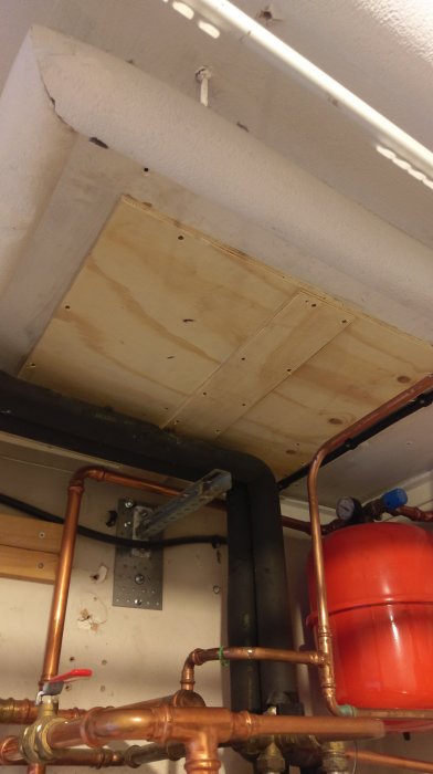 Träpanel täcker öppningen i ett tak, synliga VVS-rör och isolering i ett pannrum.