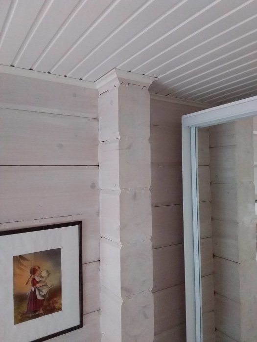 Hörn av ett rum med laminerade timmerväggar och vit takpanel, plus detalj av murad mellanvägg.