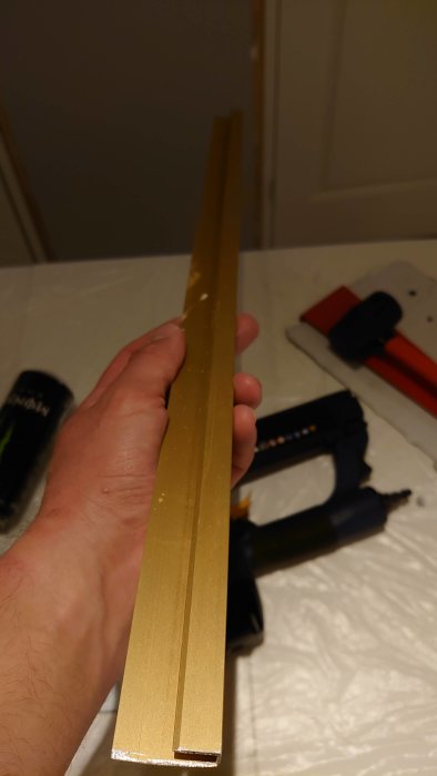 En hand håller en gyllene list framför en verktygsbänk med en häftpistol och ett verktyg.