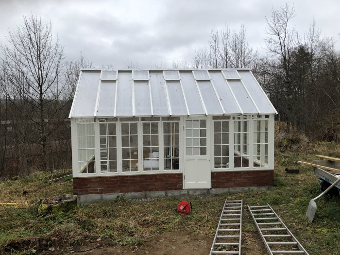 Nytt växthus med vit stomme och transparent tak mot en skogsbakgrund, murad sockel, byggmaterial i förgrunden.