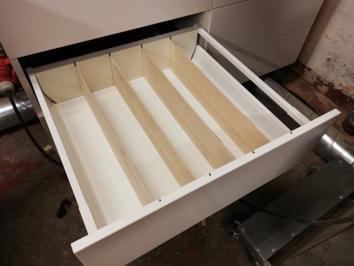 Öppen vit byrålåda med omonterade hemmagjorda träavdelare av 4mm plywood för organisering, med verktyg i bakgrunden.