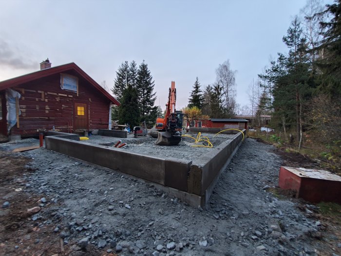 Grundarbete på kuperad tomt med balkar, makadam, byggmaskin och röd timmerstuga i bakgrunden.