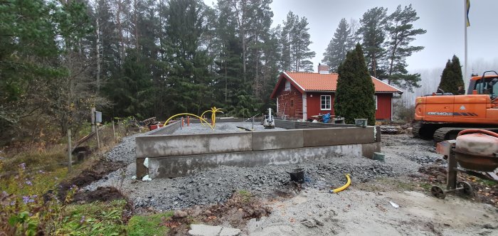 Byggarbetsplats med en ny fundament och balkar, grus och en grävmaskin framför ett rött hus.