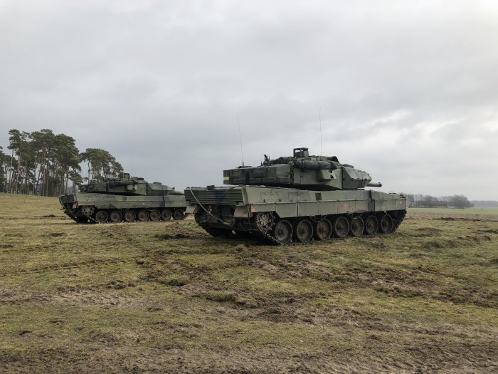 Två militära stridsfordon på övningsfält med träd i bakgrunden.