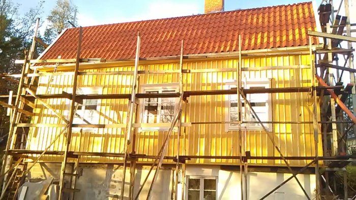 Byggnadsställningar framför en husfasad under renovering med ny panel och röda takpannor i soligt väder.