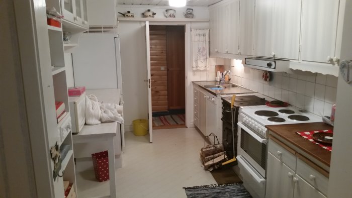 Långsmalt gammalt kök med vitt skåp, spis och öppen dörr som leder till annat rum.