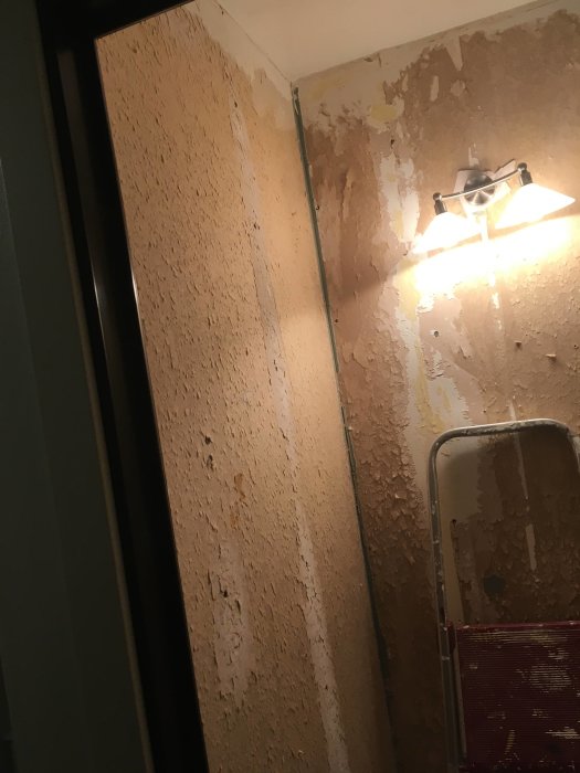 Skadad gipsvägg med rester av våtrumstapet och matta efter rivning i en gästtoalett med en stege och badrumslampa.