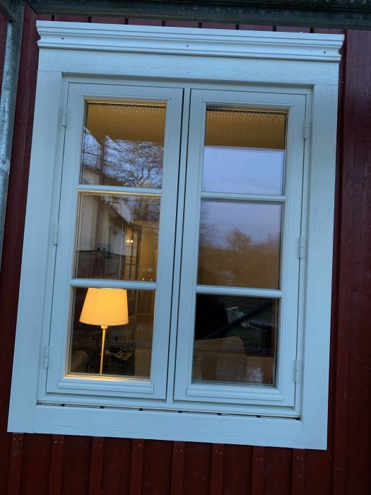 Dubbelfönster med vita karmar och spröjs, ovanför ett rött trähus, med en synlig felaktig överdel som ska bytas.