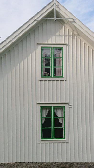 Vit träfasad med två grönmålade fönster på ett lantligt hus, övre mindre än det nedre.