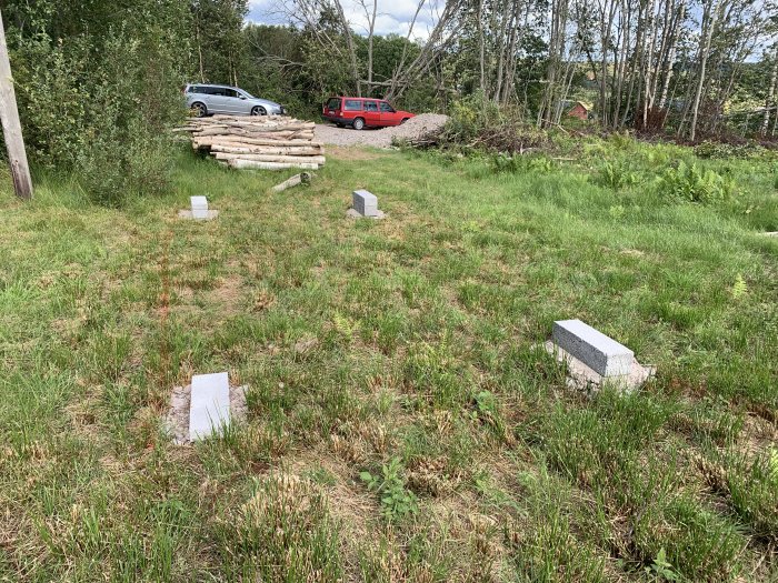 Tomtmark med stenfundament för friggebod, vedhög i bakgrunden och två bilar parkerade i periferin.