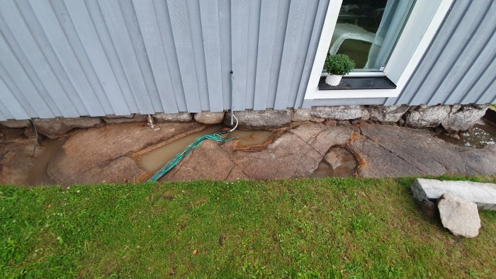 Stående vatten i djupa gropar vid en husgrund med sten och gräslighet.