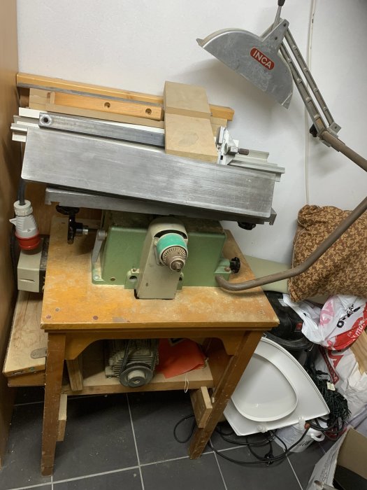En arbetsbänk med en bordsmonterad snickarmaskin och diverse trämaterial och verktyg i ett trångt utrymme.