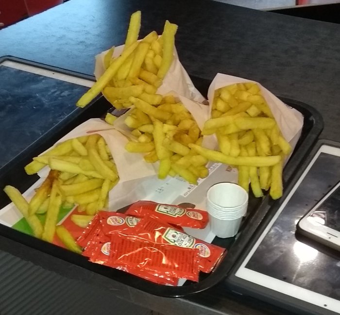 Tre portioner pommes frites på en bricka med ketchup-påsar och en smartphone.