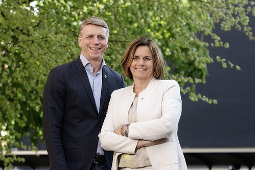 Två personer ler mot kameran, man i blå kostym och kvinna i vit jacka, med armarna i kors framför gröna träd.