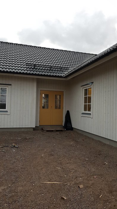 Gul entredörr på ett hus under uppbyggnad med vit fasad, gruslagd uppfart och några byggmaterial på marken.