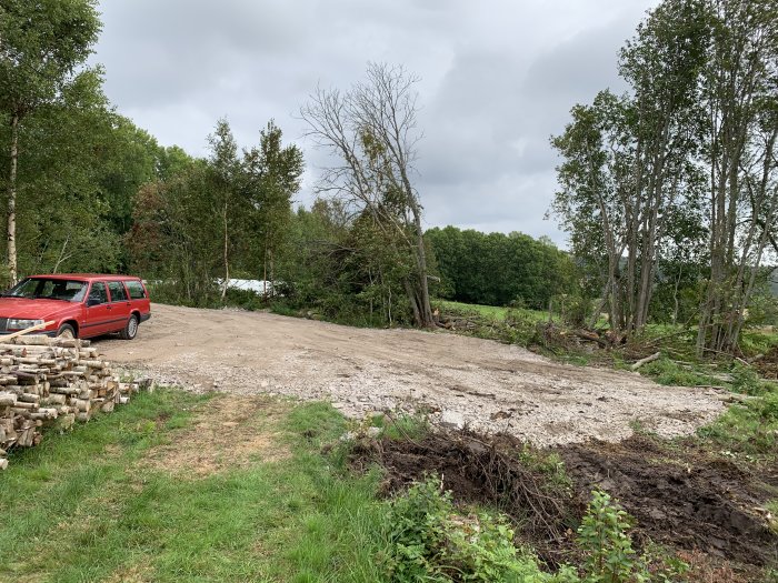 En röd bil parkerad på en nyröjd yta omgiven av nedtagen skog och vedtravar.