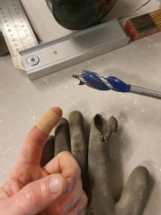 Arbetshandske med hål och drillbit samt en skadad finger med plåster, varningsbild för att inte borra med handskar.