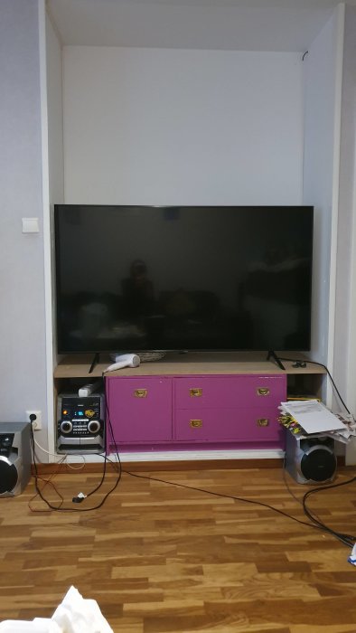 En 65 tum stor TV på en lila låg möbel med gula lådor i ett rum med parkettgolv, planerad plats för TV-möbelbygge.