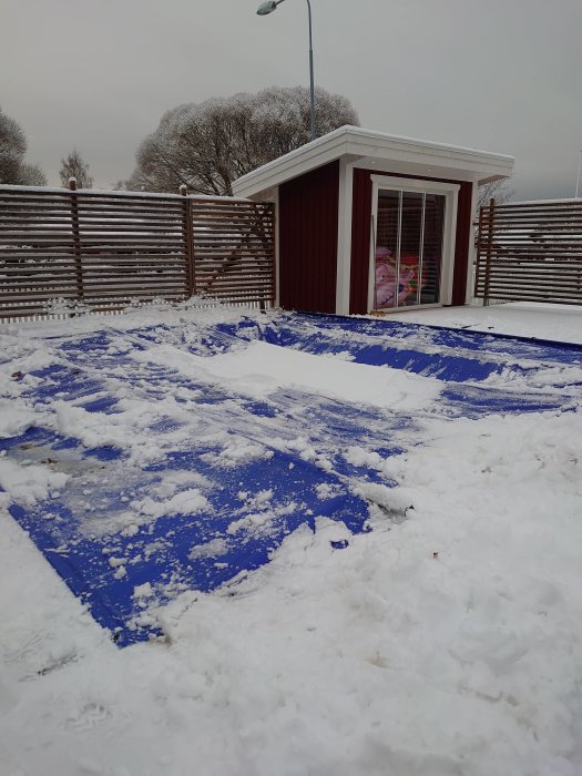 Skyddstäckt poolområde med snö och trädgårdsbod i bakgrunden under en vinterdag.