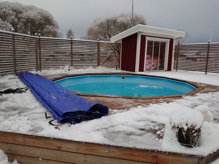 Pool med delvis upprullat blått skydd på snötäckt träaltan och vinterklädd vegetation i bakgrunden.