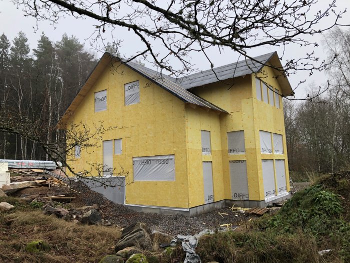 Hus under konstruktion med gul isolering och temporärt täckta fönsteröppningar.
