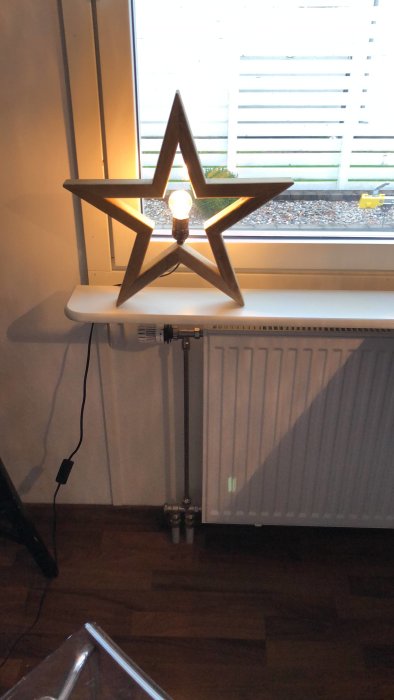 Trästjärna med tänd glödlampa framför fönster på vit hylla ovanför radiator.