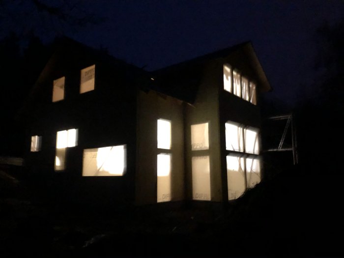 Kvällsbild av ett hus under konstruktion med tända lampor inomhus och mörk himmel i bakgrunden.