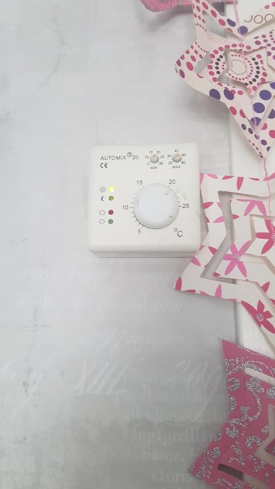 Termostat inställd på cirka 21 grader med indikatorer på vit vägg, bredvid rosa och lila papperspynt.