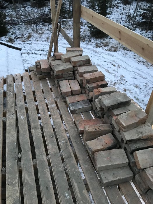 Stapel med gamla tegelstenar på en träställning, snöigt landskap i bakgrunden, indikerar rivet skorstensprojekt.