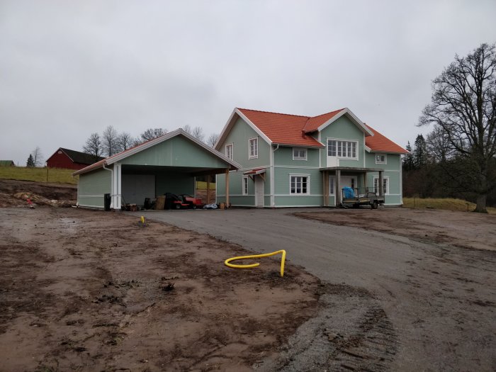 Nybyggt ljusgrönt hus med rött tak, carport och grusad infart under uppbyggnad, omgiven av en lerig tomt.