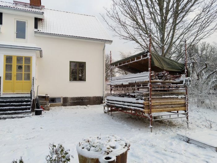 Ställning täckt med presenning på snöig mark bredvid hus med gul dörr.